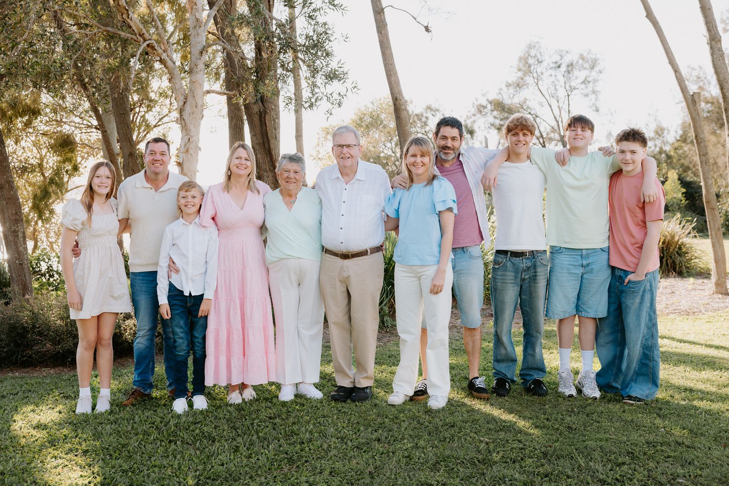 Large multigenerational family smiling together outdoors on green grass surrounded by tall trees.