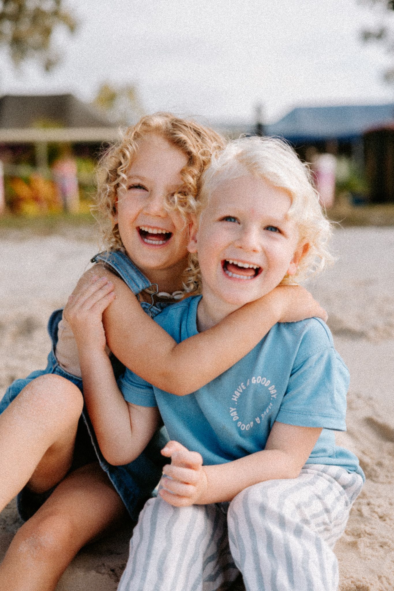 Two laughing blonde children hugging on sandy beach, wearing blue outfits, sharing a joyful moment outdoors.