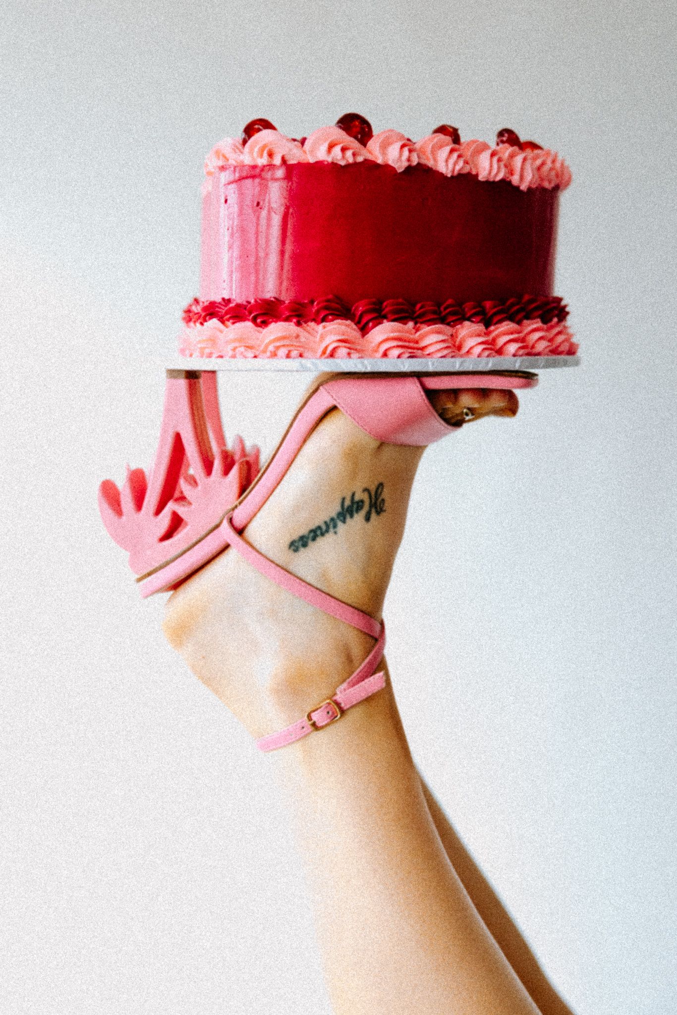 Hand holding a red and pink layered cake on a plate while wearing a pink high heel sandal.