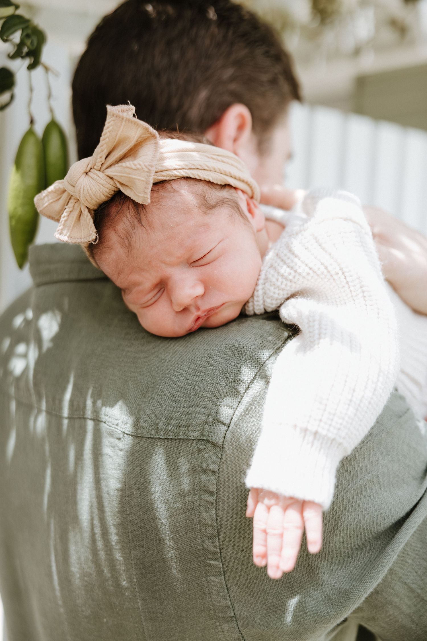Sleeping newborn baby wearing white sweater and peach bow headband rests peacefully on shoulder in soft natural light.