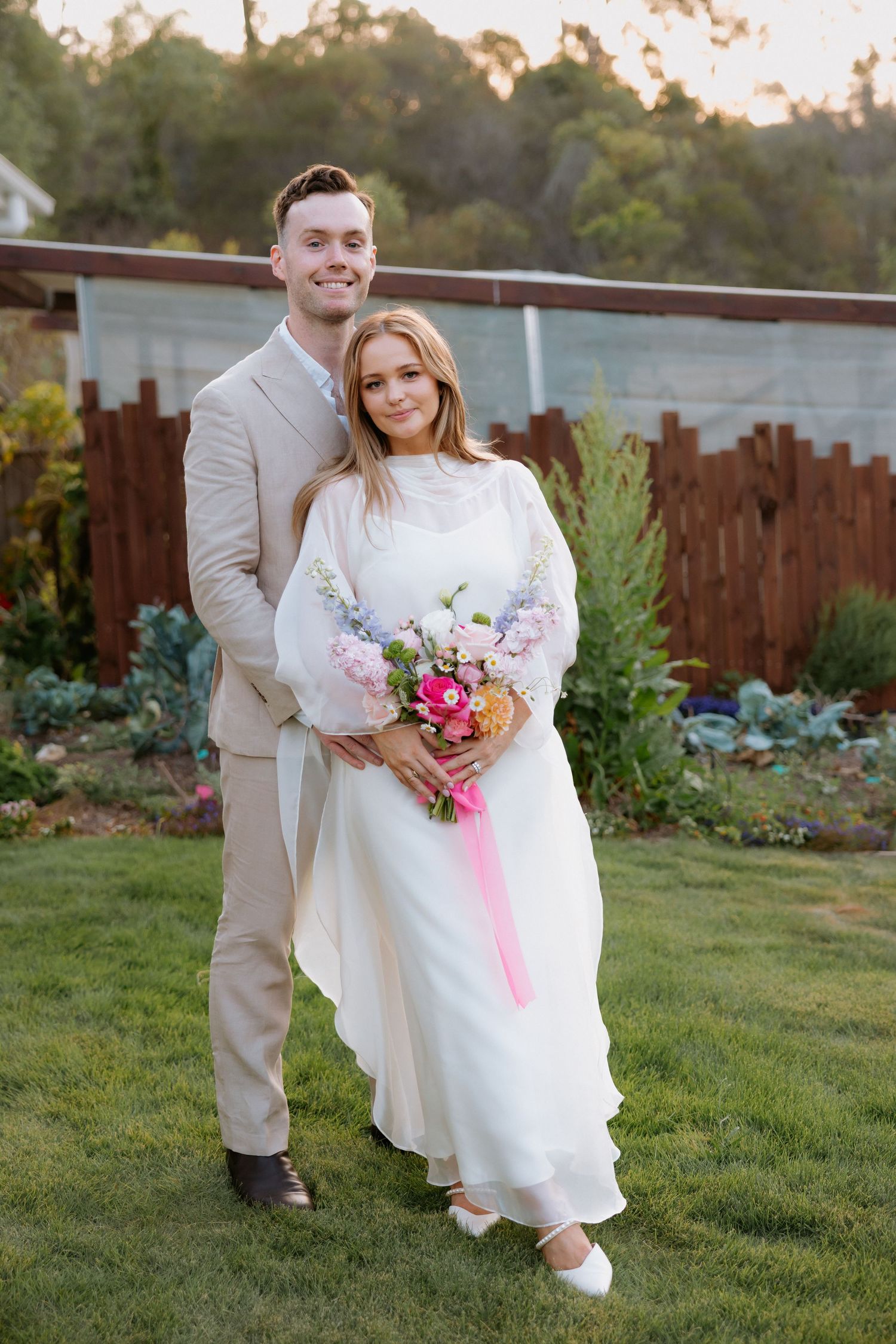Wedding couple posing in a backyard garden with wooden fence and sunset lighting.