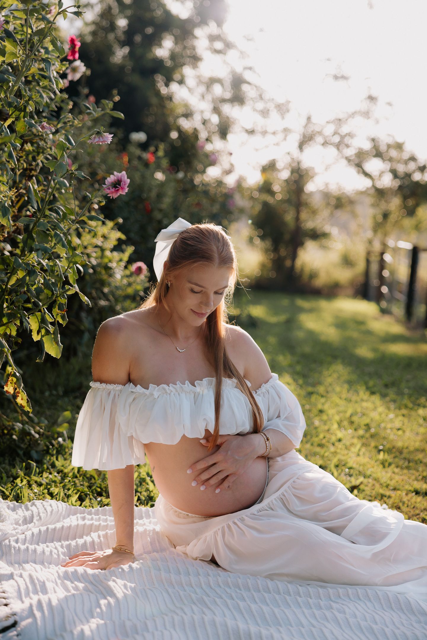 Pregnant woman in white off-shoulder dress sitting on blanket outdoors, cradling baby bump near blooming flowers in sunlight.