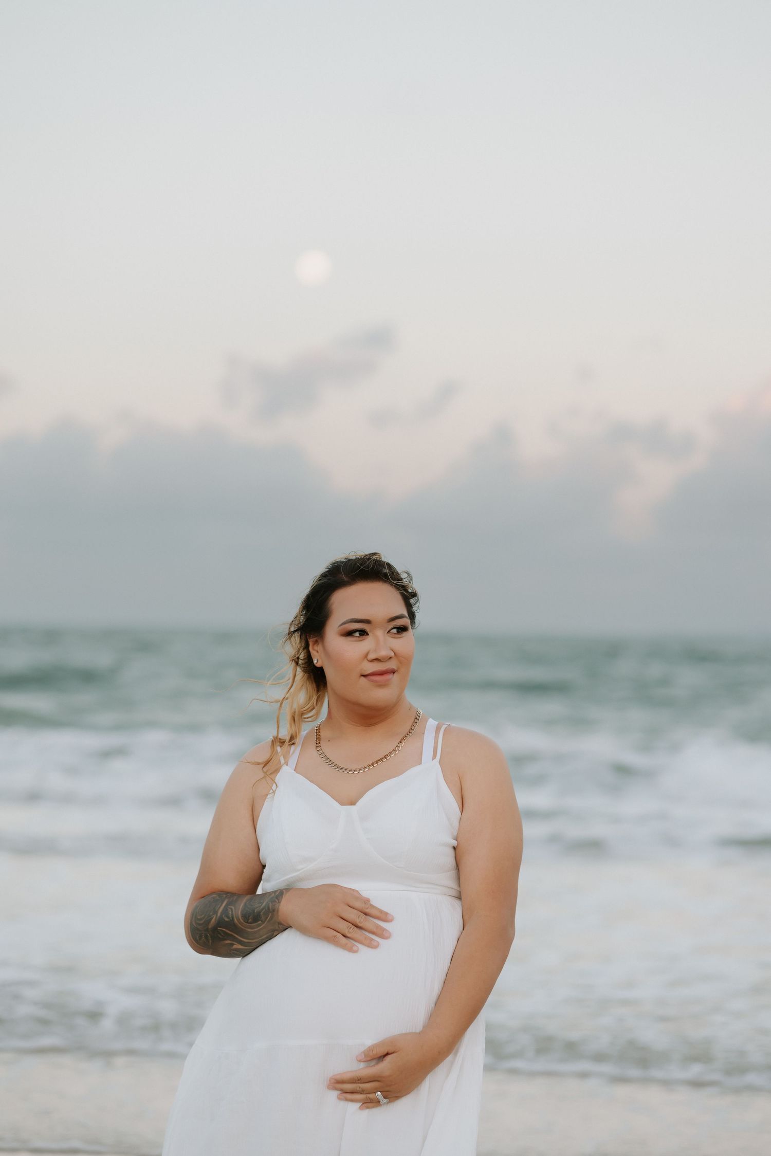 A pregnant woman in a white dress poses at sunset on a beach with waves and soft pastel sky in the background.
