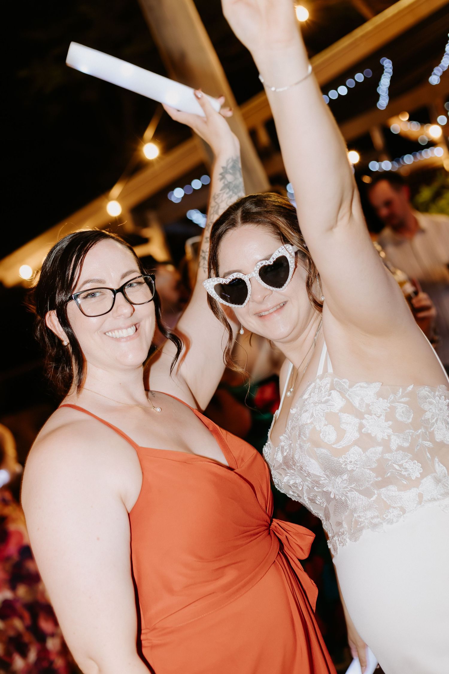 Two guests celebrating with glow sticks under string lights at evening wedding reception.