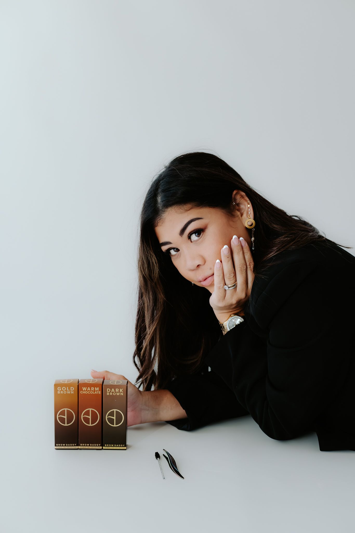 Woman in black posing with three brown makeup products on a white surface.