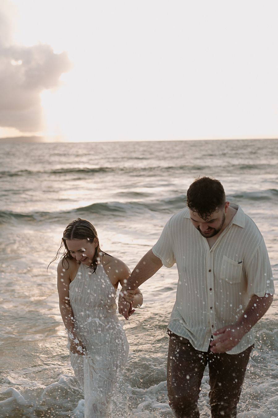Couple holding hands walking through ocean waves at sunset on a sandy beach, wearing white outfits.