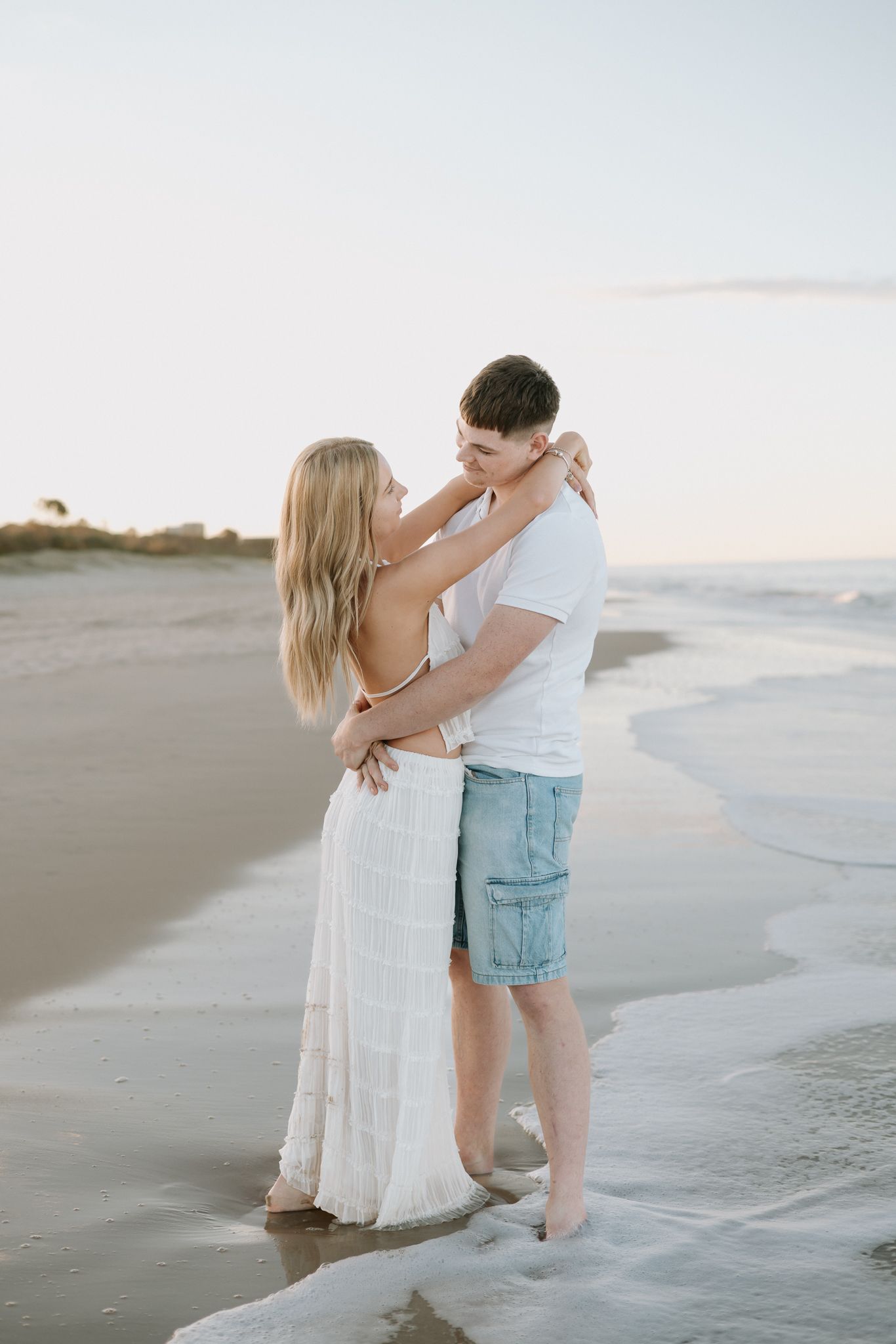 Romantic couple embracing on a sandy beach at sunset, woman in white dress and man in blue shorts.