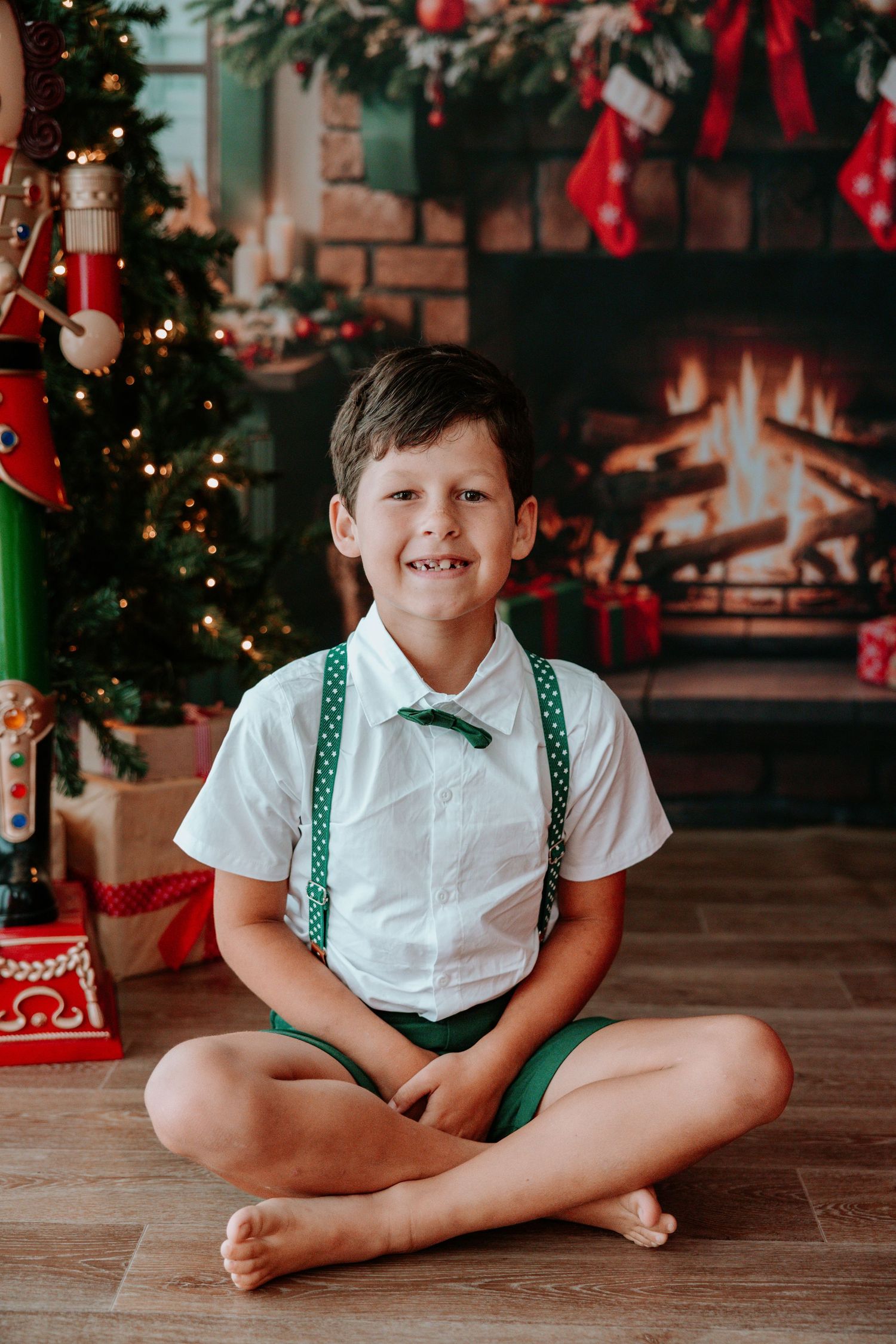 Smiling boy in white shirt and green suspenders sitting cross-legged by a decorated Christmas tree and fireplace.