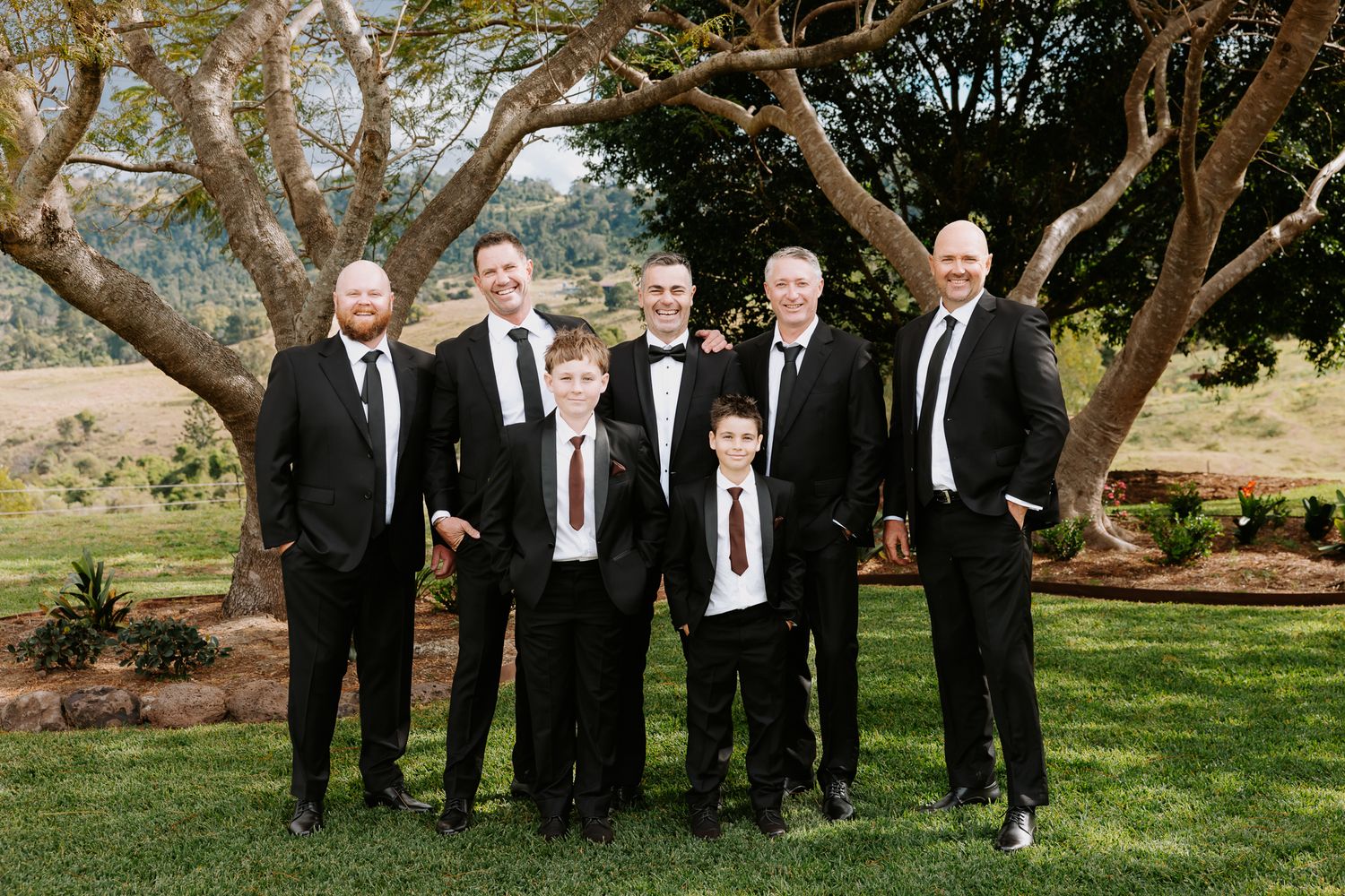 Group of men and boys in black suits posing outdoors on green lawn with trees and scenic landscape background.