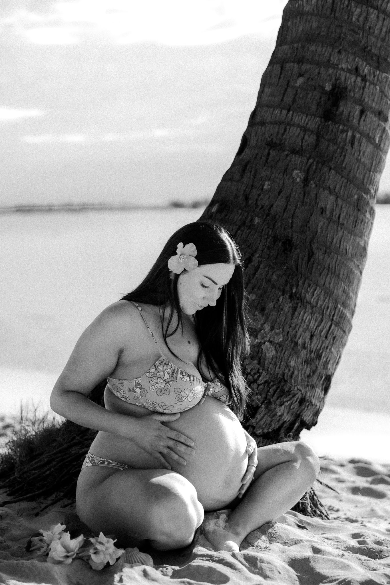 Black and white photo of a pregnant woman in a bikini sitting cross-legged on a beach, cradling her belly near a palm tree.