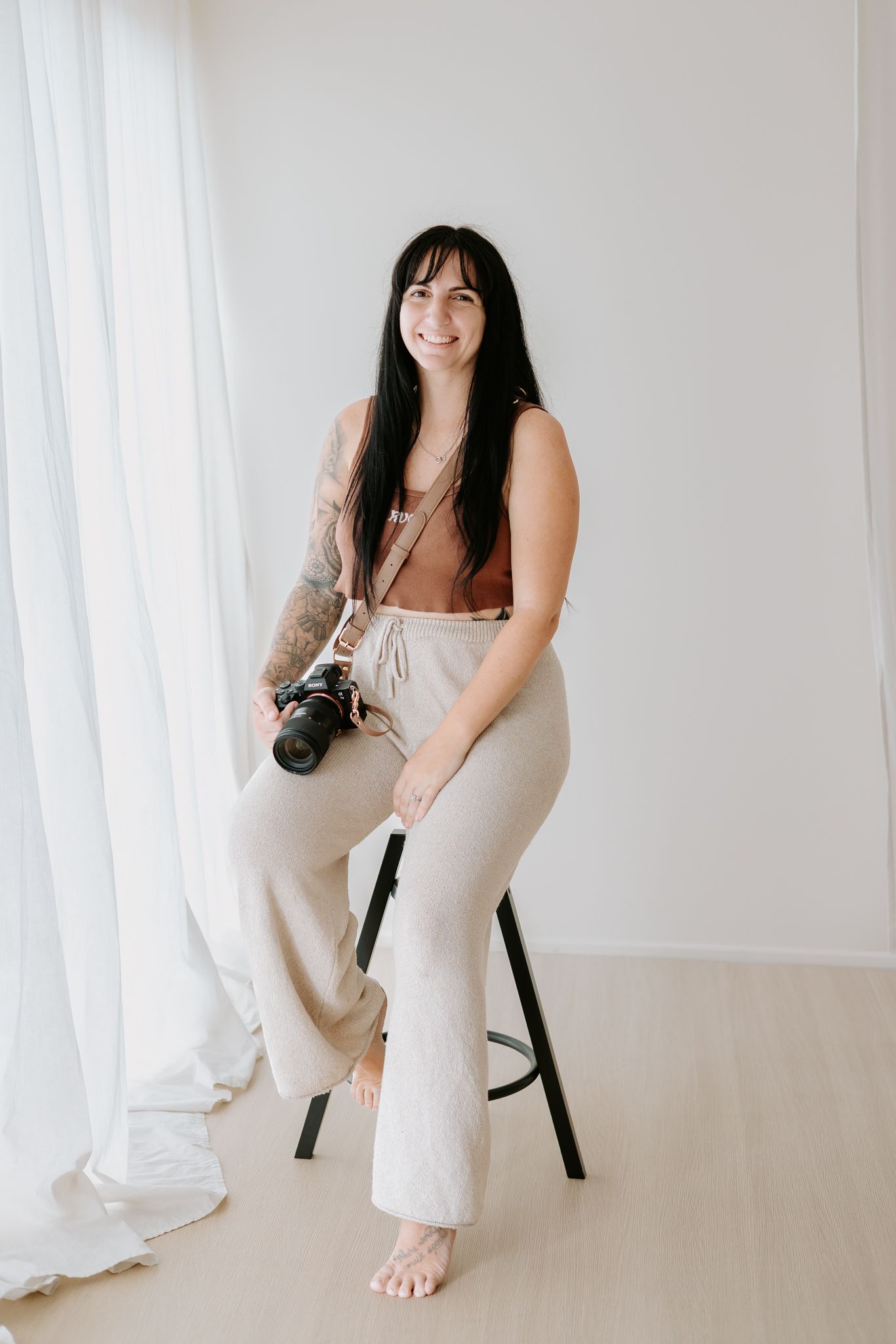 Smiling woman with dark hair sits on a stool holding a camera, wearing beige pants and a brown top in a bright studio.