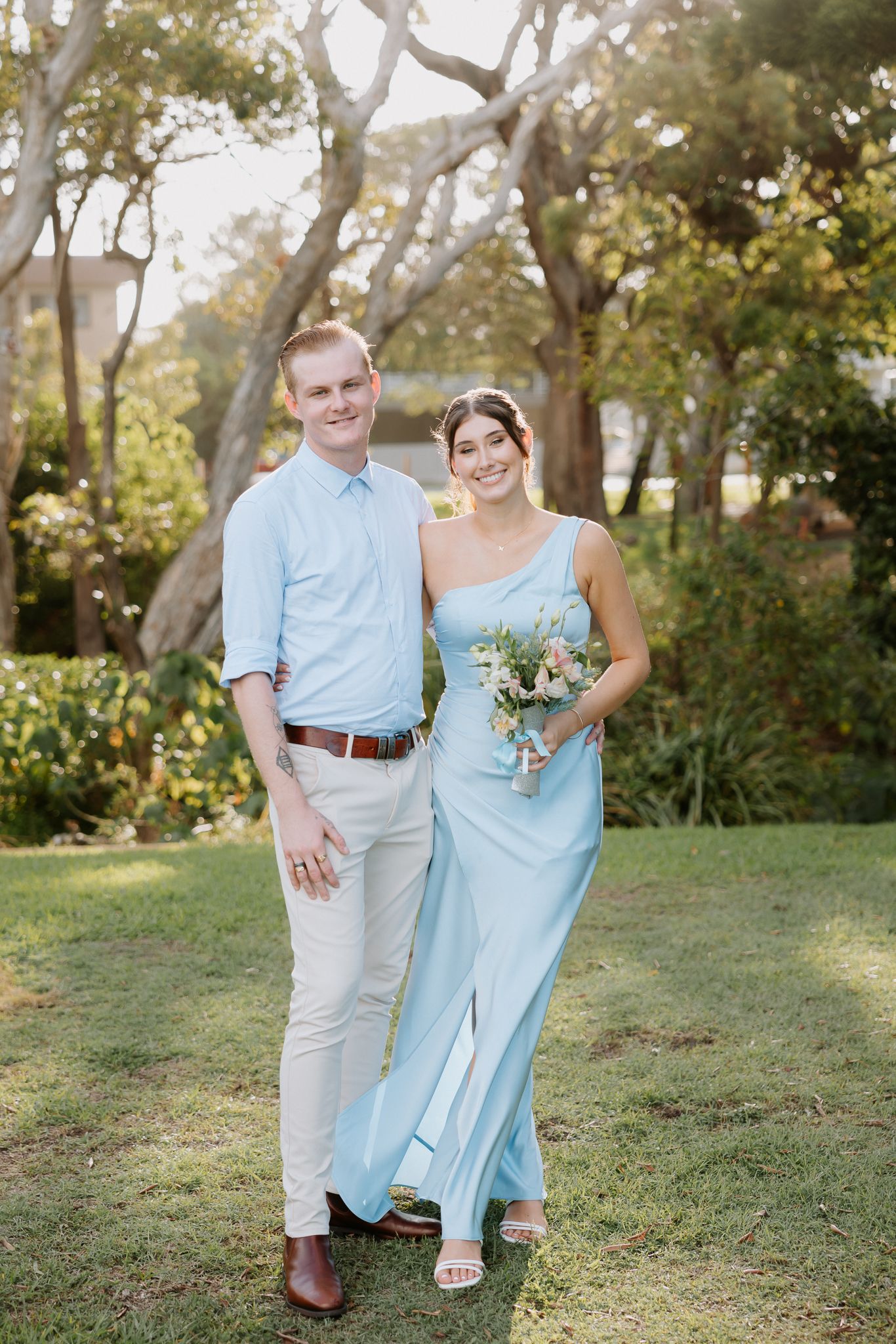 Couple in matching light blue outfits posing outdoors, bride holding bouquet, surrounded by lush greenery.