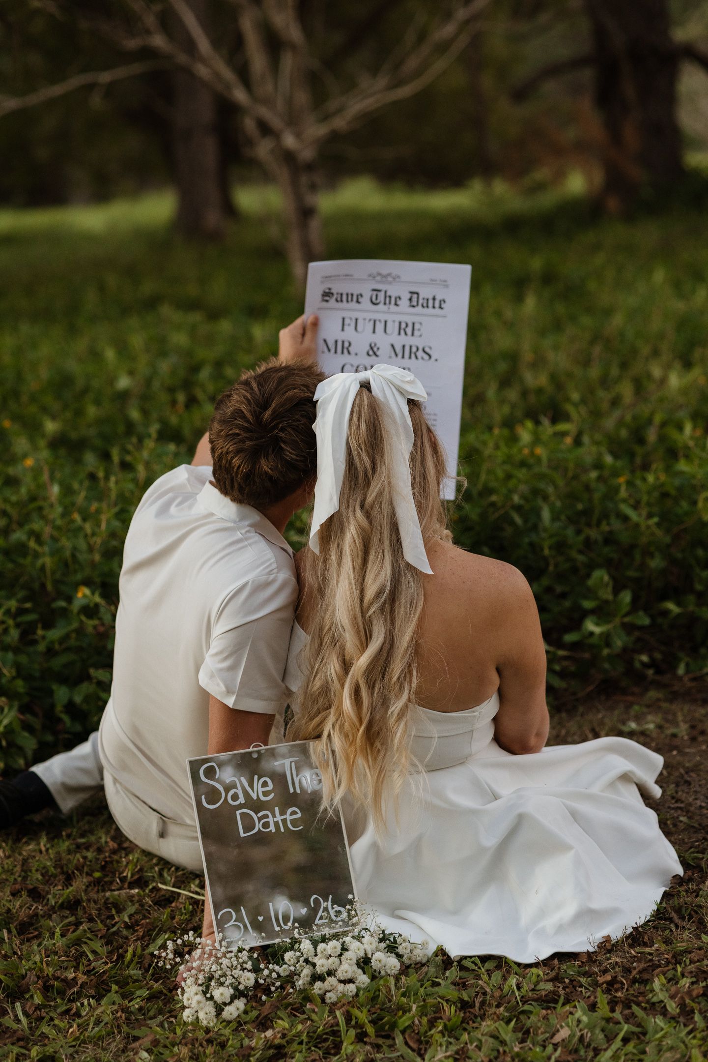 Couple sitting on grass holding Save the Date wedding announcement signs, bride in white dress with long blonde hair.
