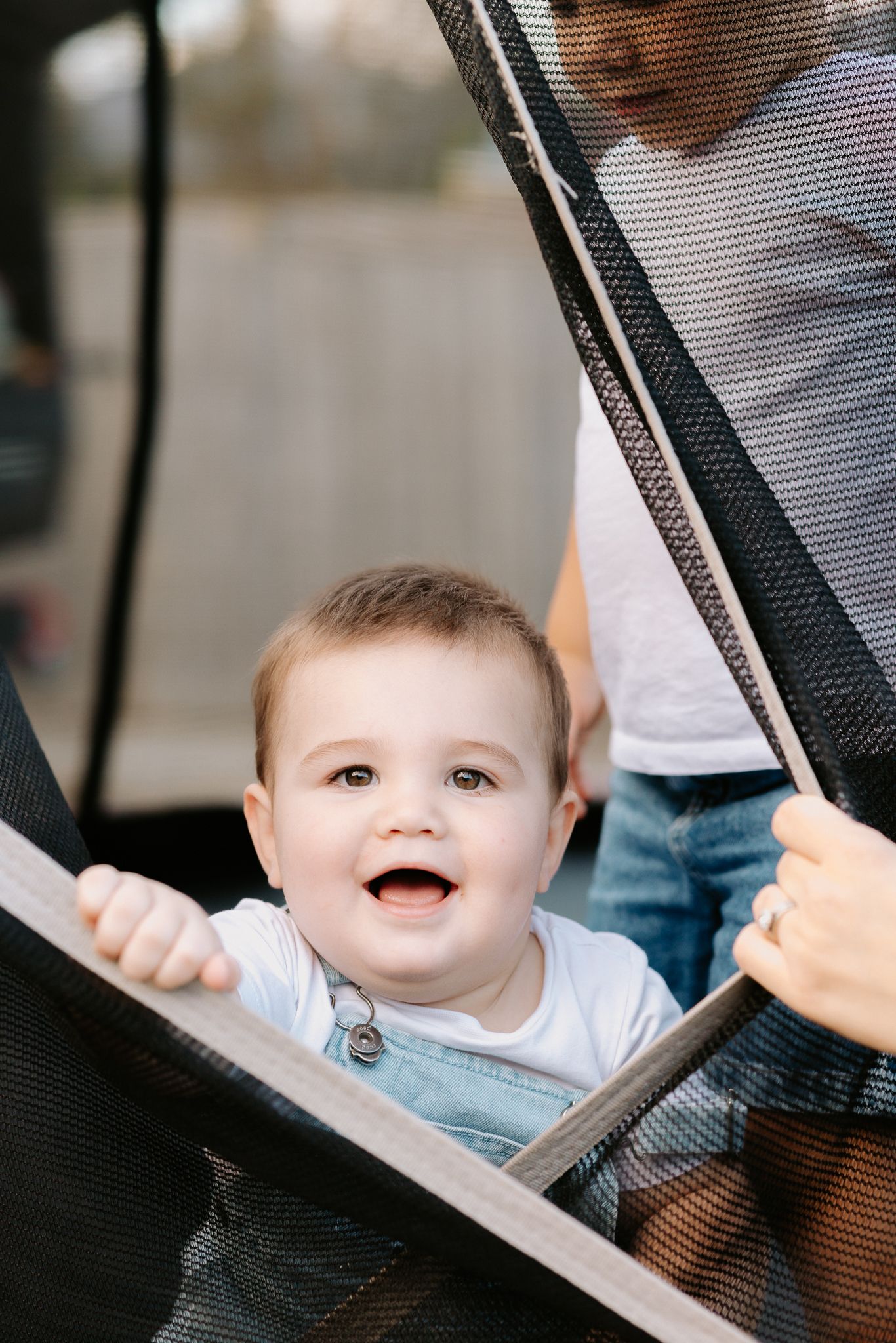 Happy baby in overalls smiles from stroller while caregiver opens mesh side panel outdoors.