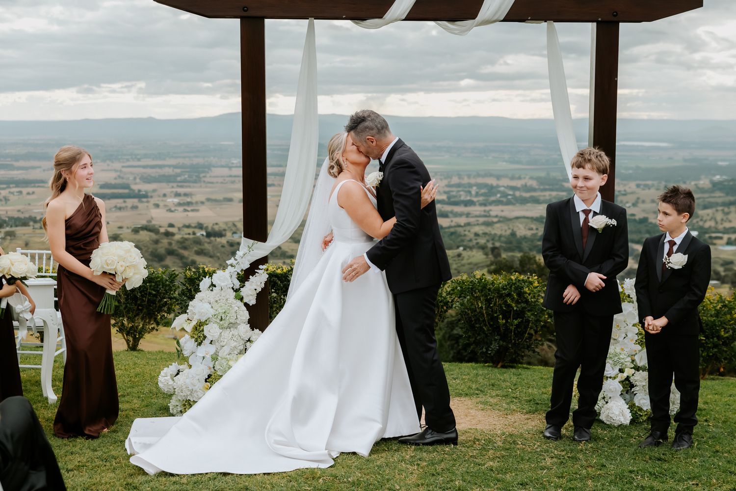 Bride and groom share first kiss at outdoor wedding ceremony with scenic valley views and white floral arch.