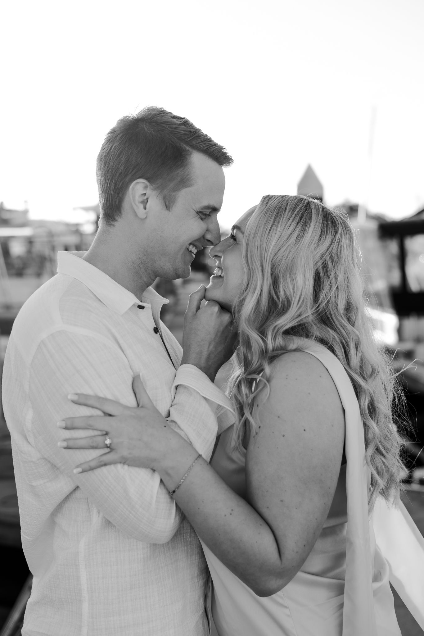 Black and white photo of a smiling couple embracing and about to kiss outdoors, woman wearing engagement ring.