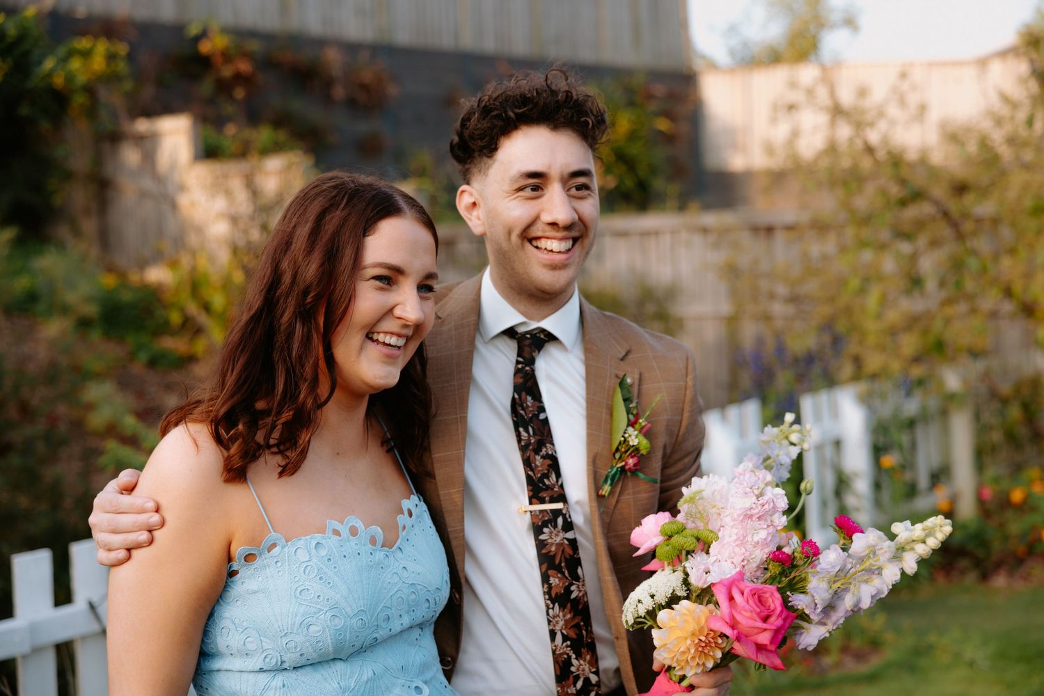 Smiling couple in wedding attire, man in brown suit with floral tie, woman in blue dress holding bouquet outdoors.