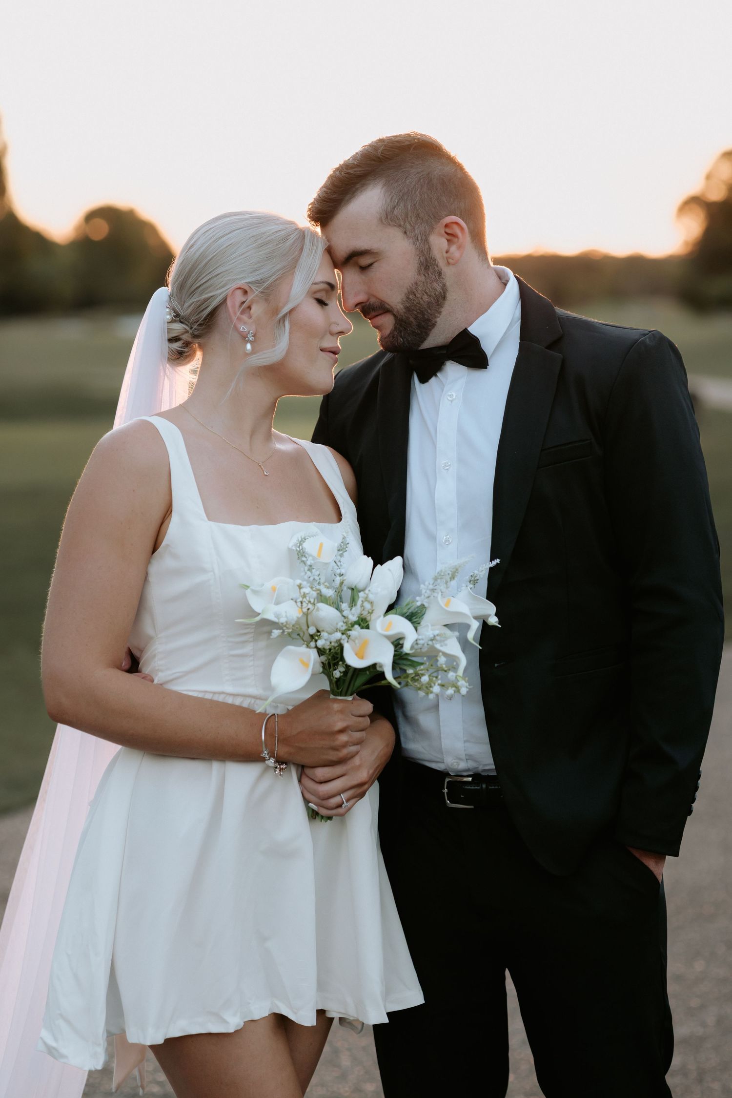 Bride in short white dress holding calla lily bouquet with groom in black tuxedo at golden hour outdoor wedding.