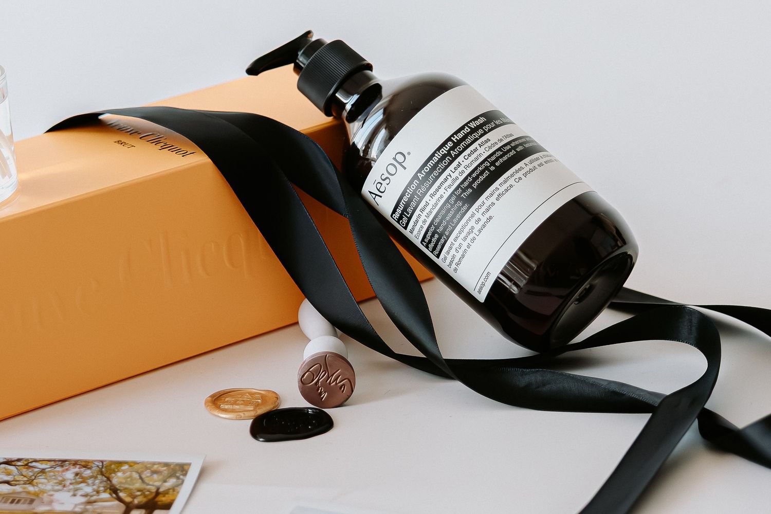 Aesop skincare bottle with black ribbon on white surface beside orange gift box and wax seal coins.