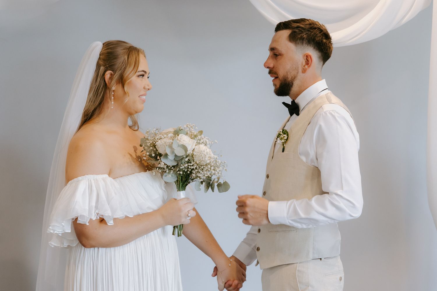 Bride in white off-shoulder gown holding bouquet exchanges vows with groom in beige suit at wedding ceremony.