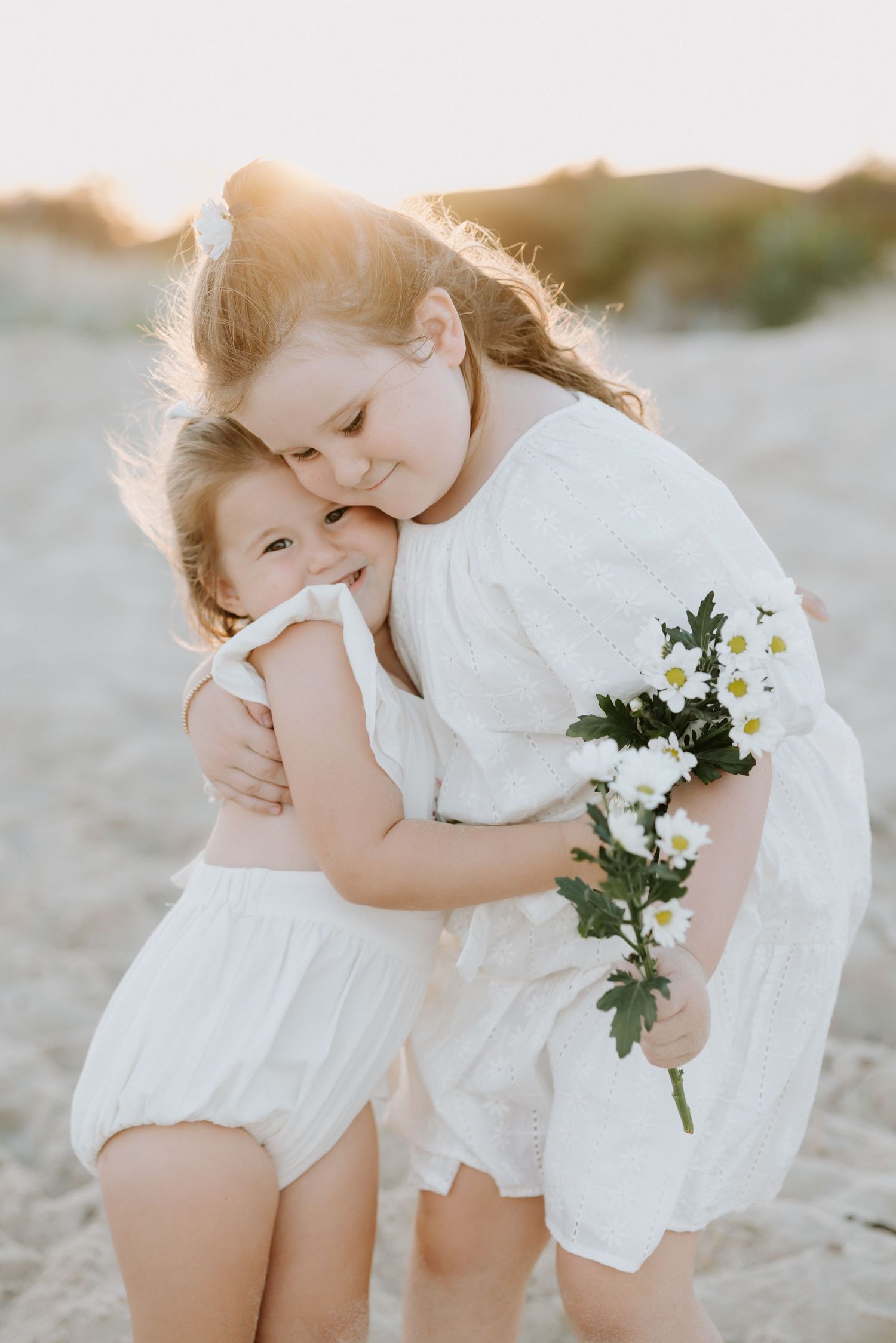 Two figures in white clothing embrace tenderly on a sandy beach at sunset with white flowers in the background.