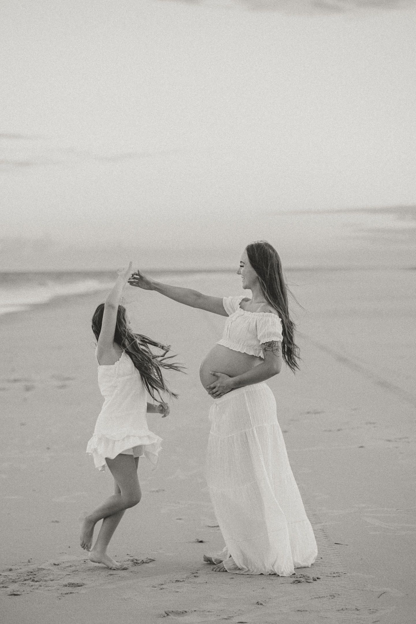 Two people in white dresses twirling and playing together on a sandy beach in black and white photography.