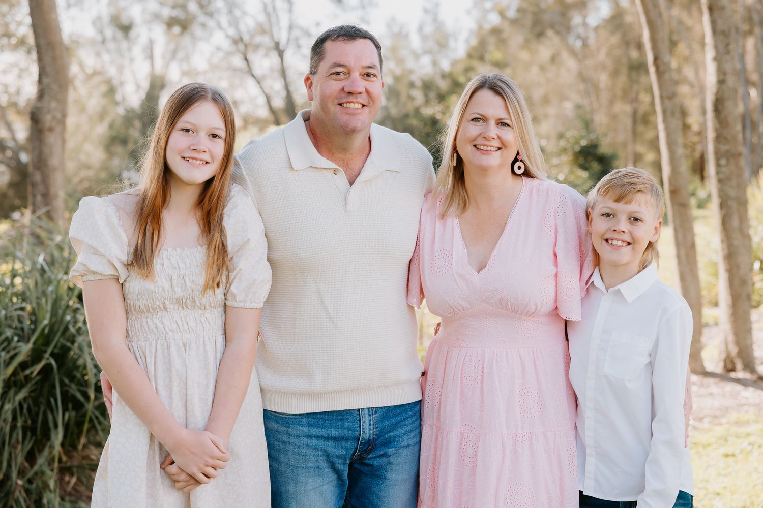 Happy family of four smiling outdoors in casual attire, posing together in a sunny park setting.