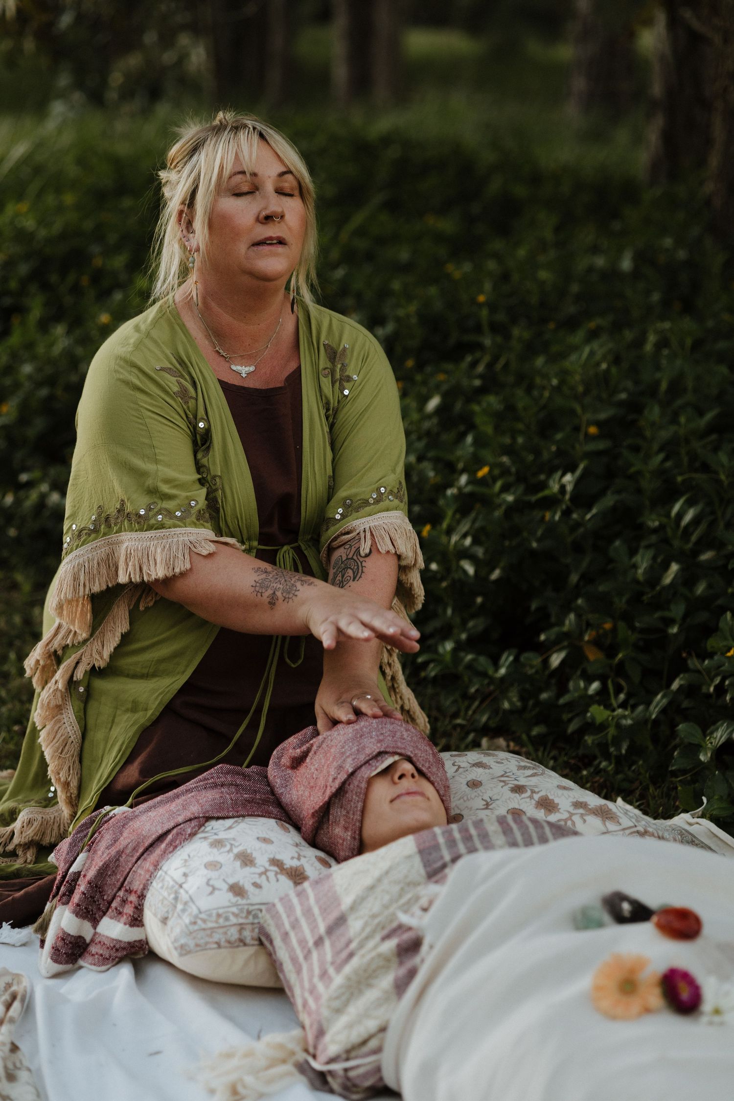 Woman in green kimono performing outdoor energy healing session on person lying on white blanket surrounded by nature.