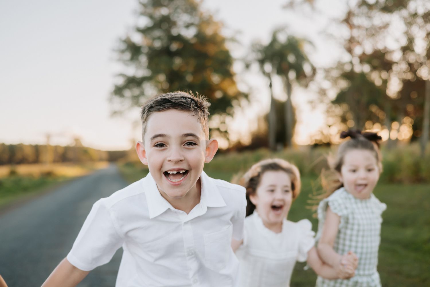 Three happy children running outdoors on a rural path during golden hour, smiling and laughing in a joyful moment.