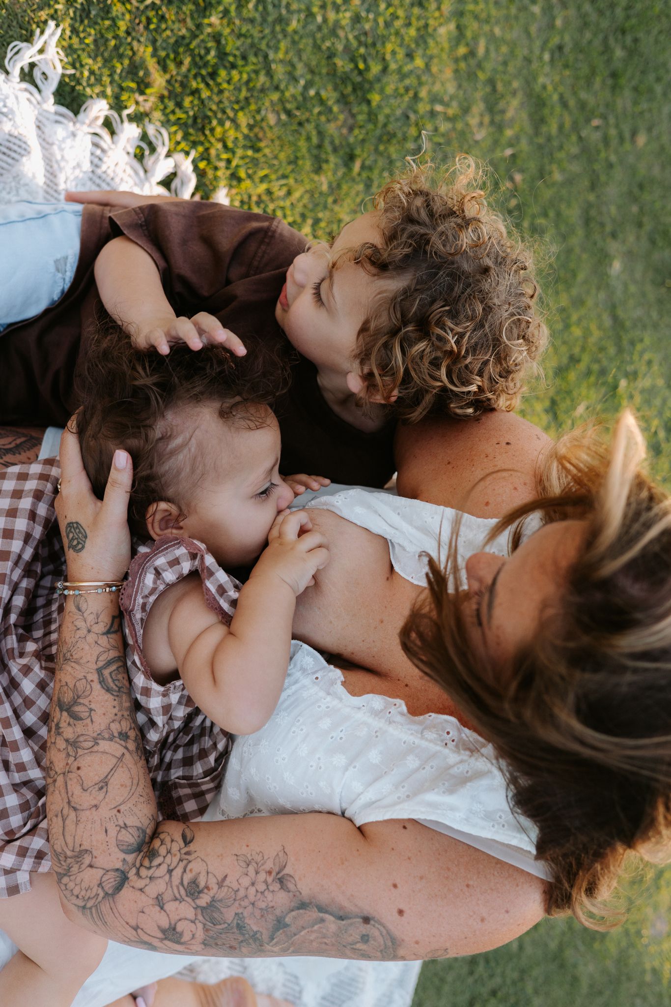 A tattooed mother breastfeeds her baby outdoors on grass while her curly-haired toddler kisses her cheek.