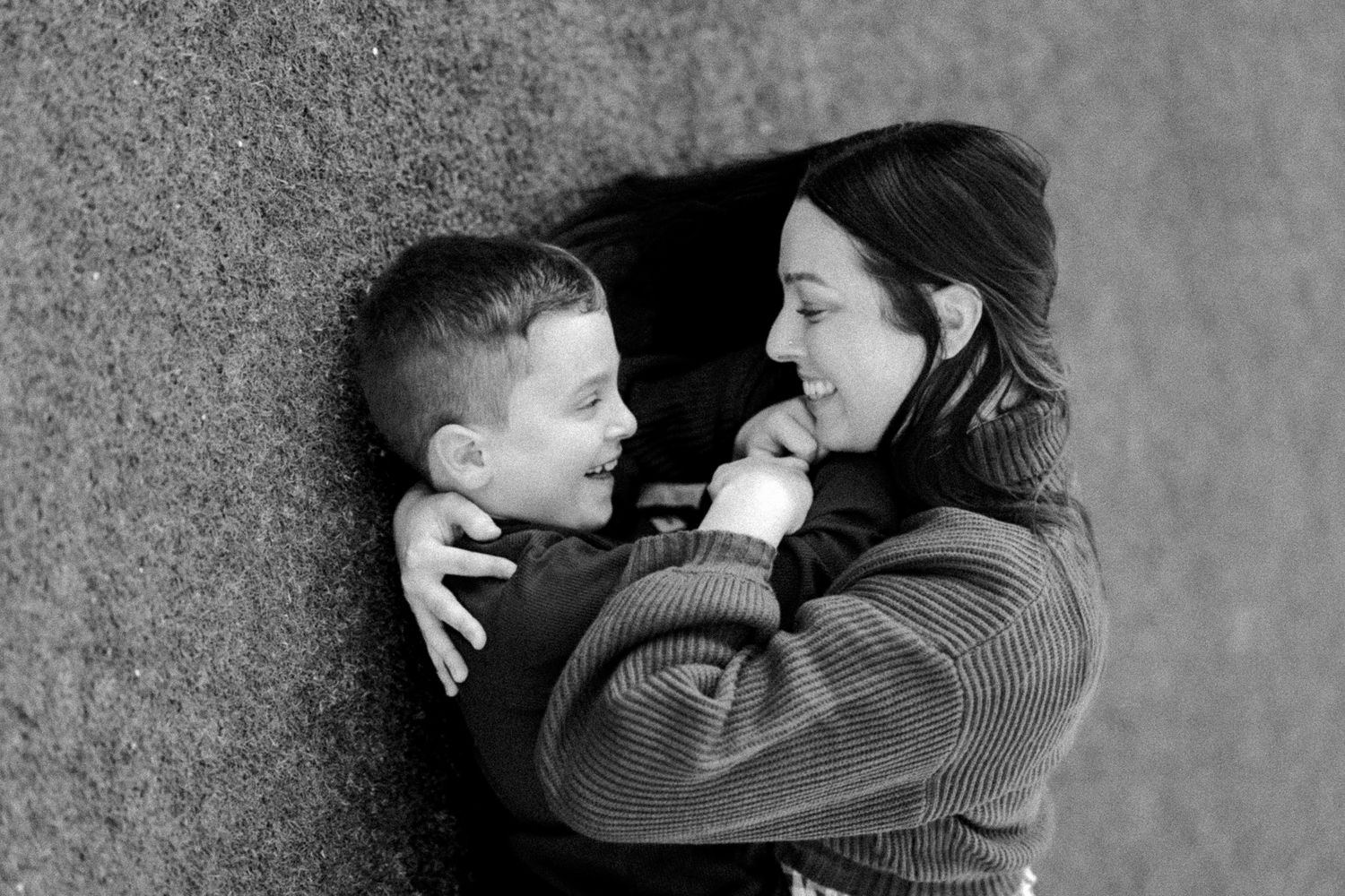Black and white photo of a smiling mother and young son sharing a tender moment against a textured wall.