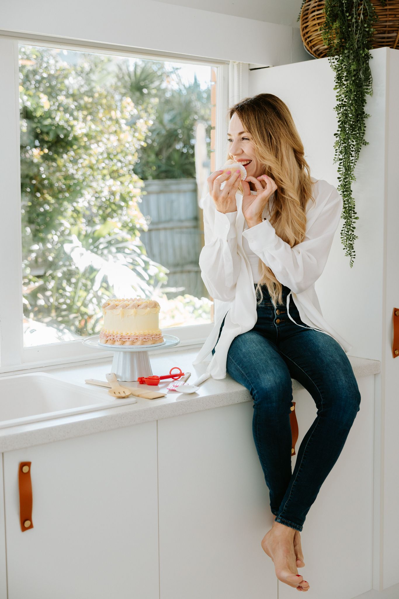 Woman in white shirt and jeans sitting on kitchen counter beside a layered cake on a stand near a bright window.