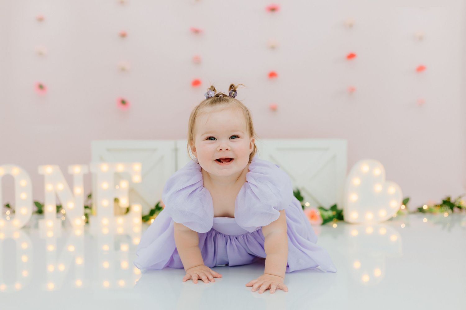 A baby in purple clothing crawls on a white surface with twinkling lights and pink decorations in the background.