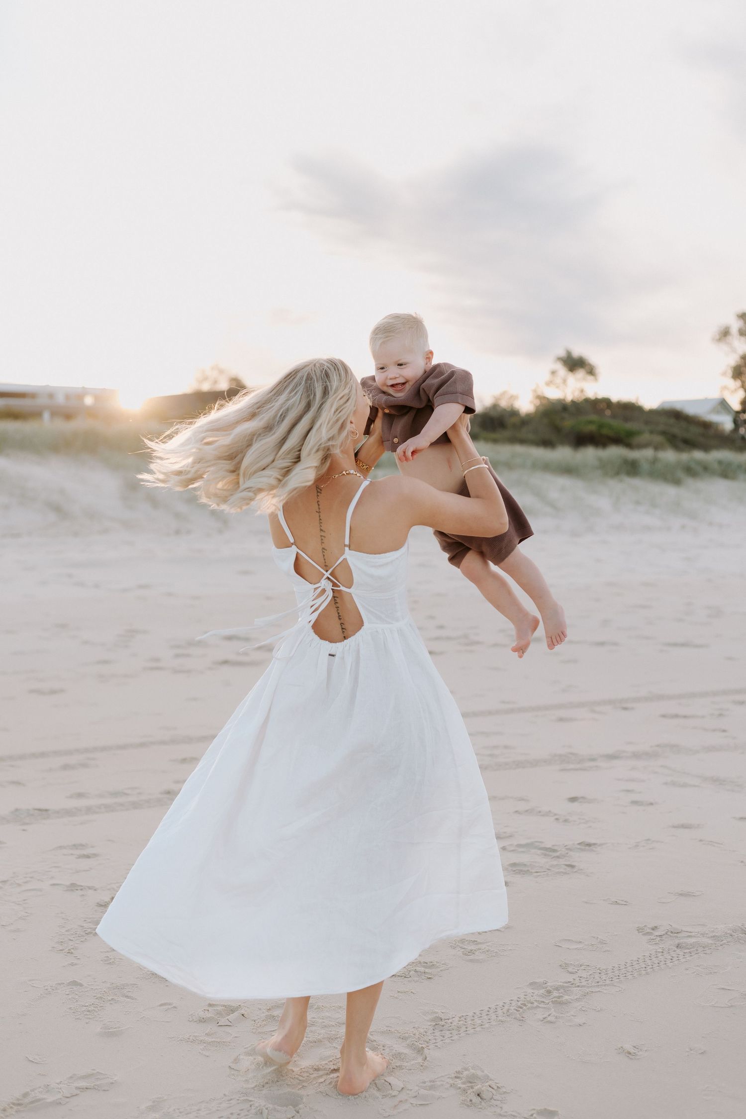 Blonde woman in white open-back dress twirling baby on sandy beach at sunset.