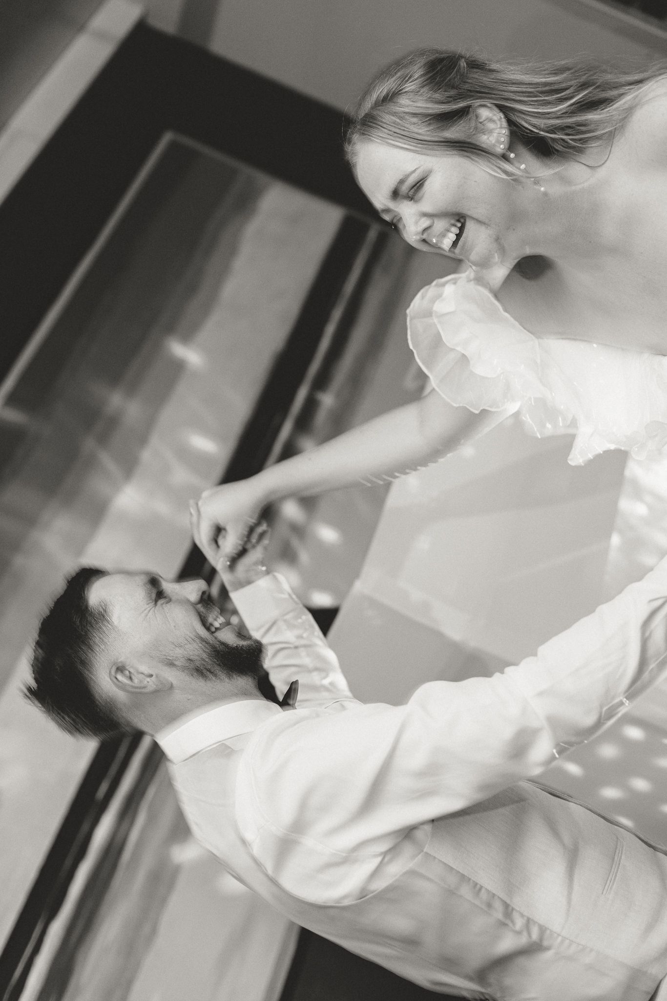 Black and white photo of a laughing bride in white dress dancing joyfully with a smiling groom in white shirt.