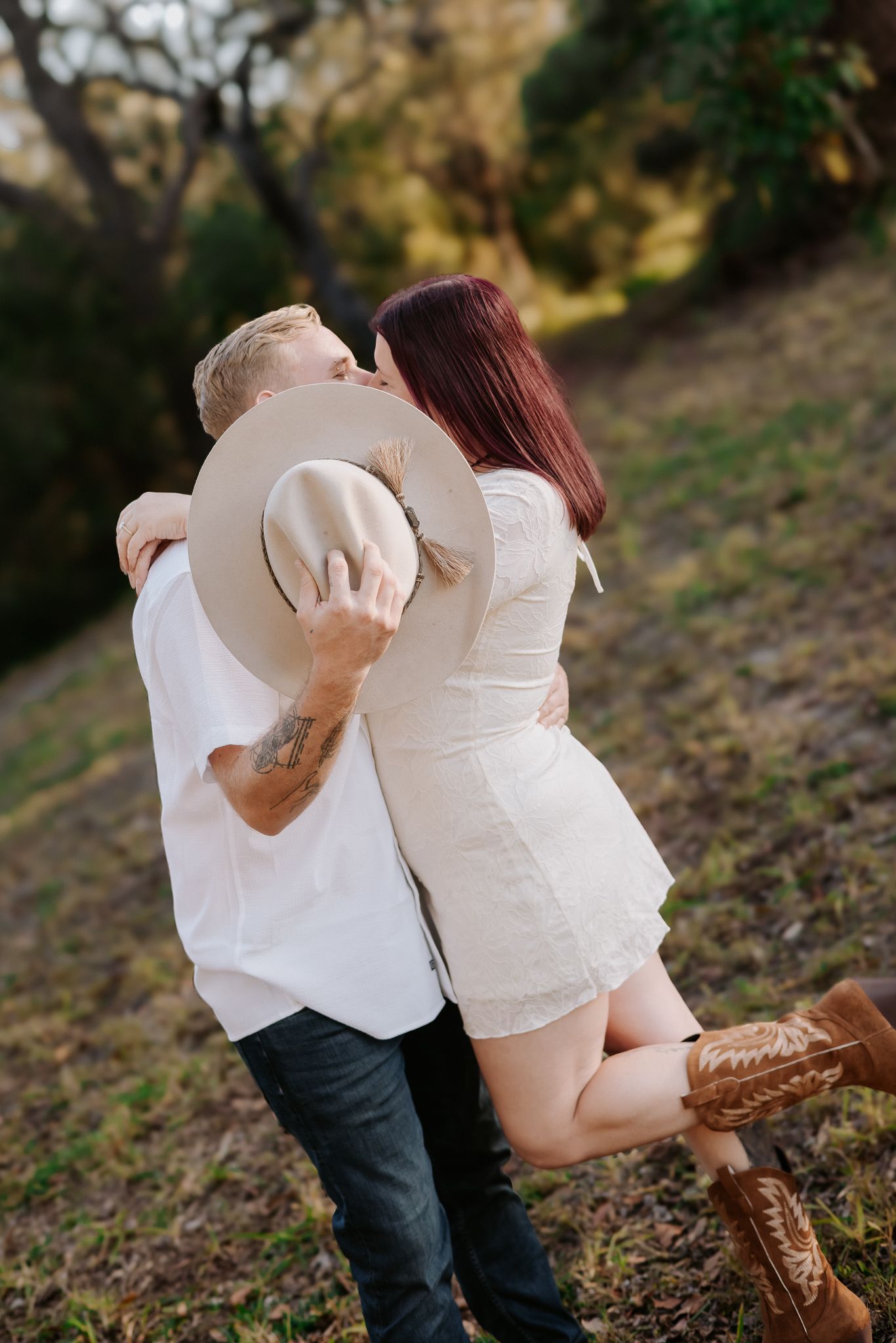 Couple sharing a romantic kiss outdoors, woman in white dress and cowboy boots lifted off ground, man in white shirt holding hat.