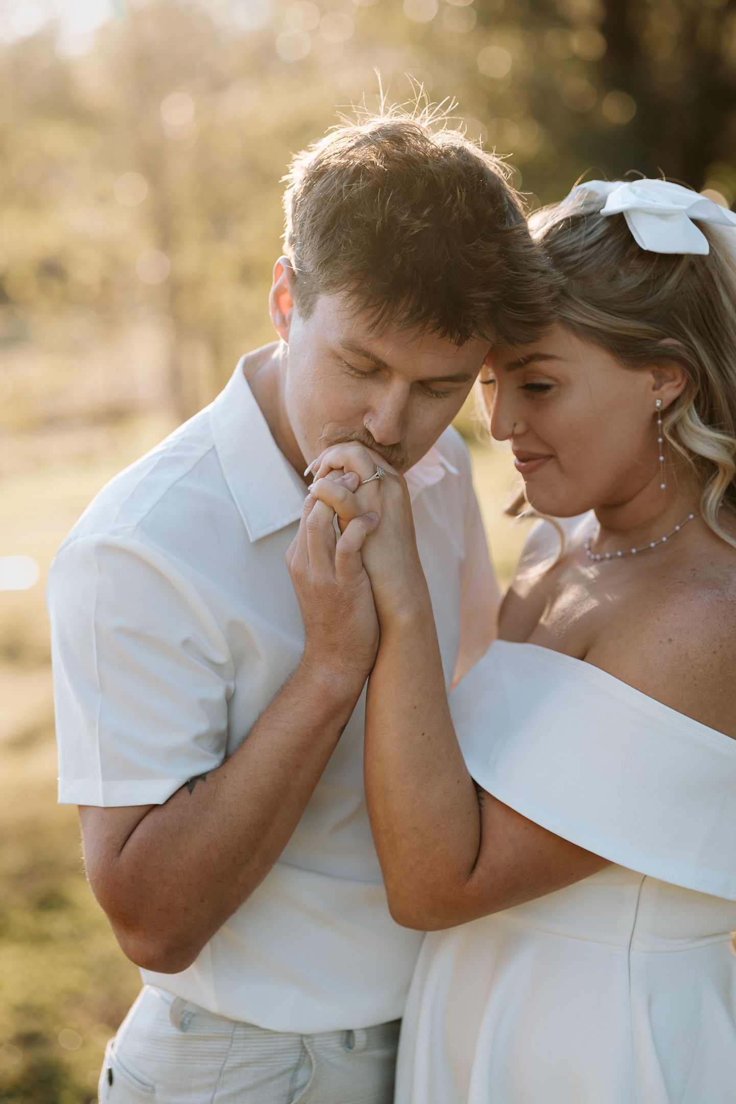 Groom kissing bride's hand during golden hour outdoor wedding portrait, bride wearing white off-shoulder gown.