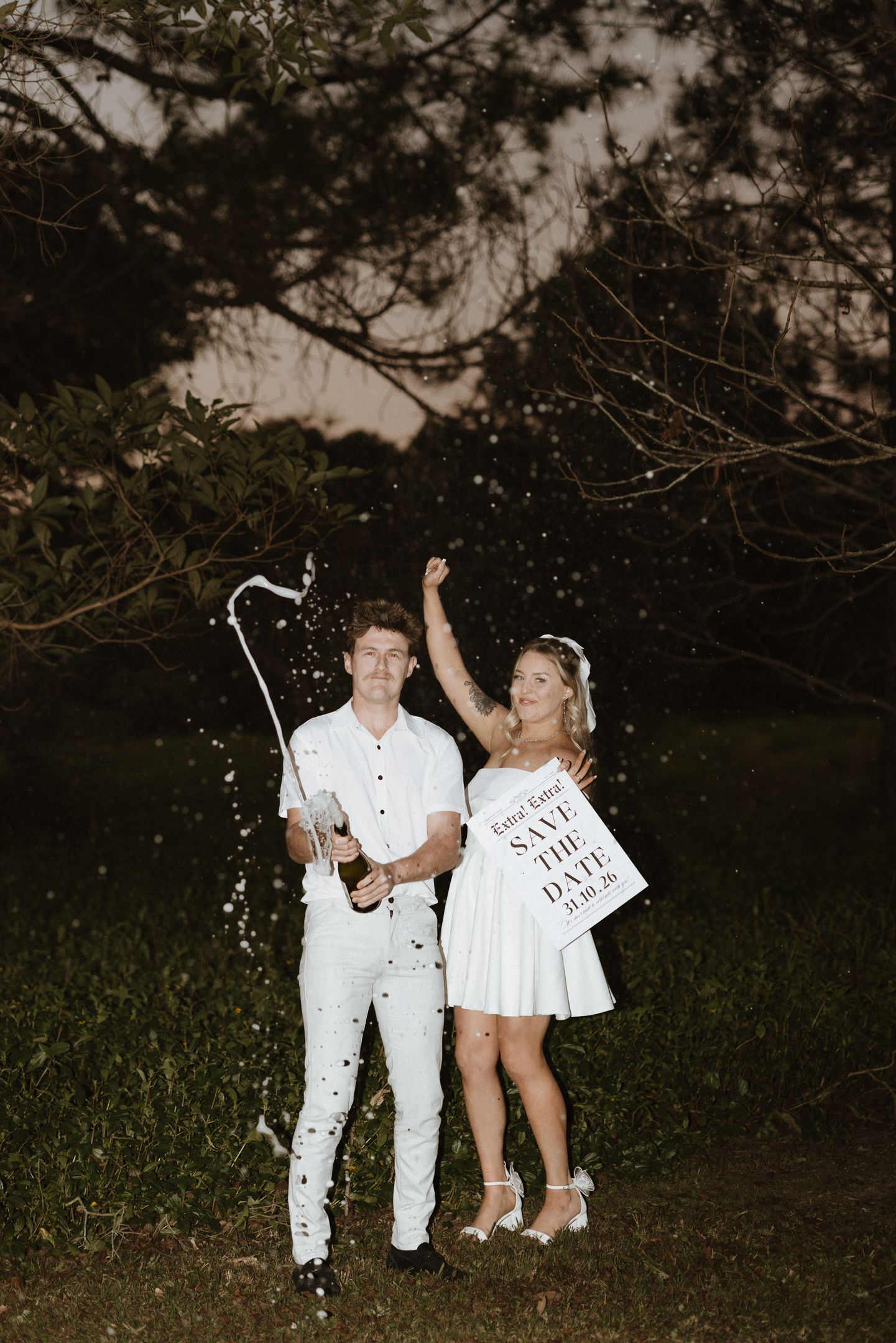 Couple in white outfits celebrating engagement, popping champagne and holding Save the Date sign outdoors at night.