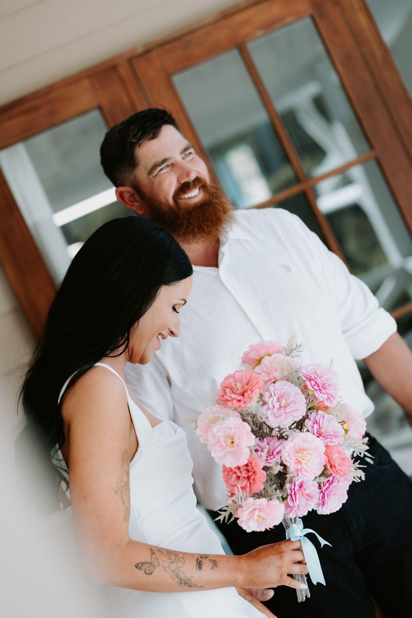 A couple embraces while holding a pink floral bouquet with carnations and baby's breath in an indoor setting.