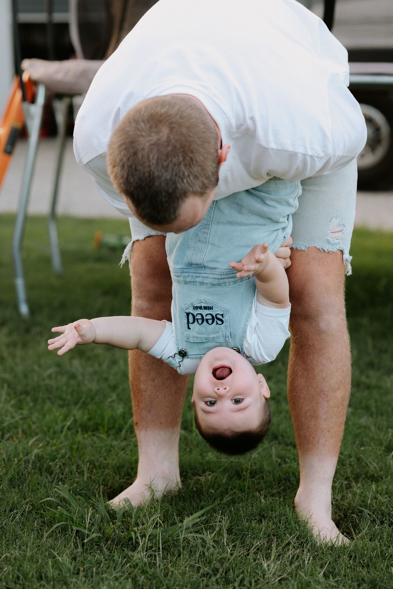 Father playfully holds laughing baby upside down on green grass outdoors.