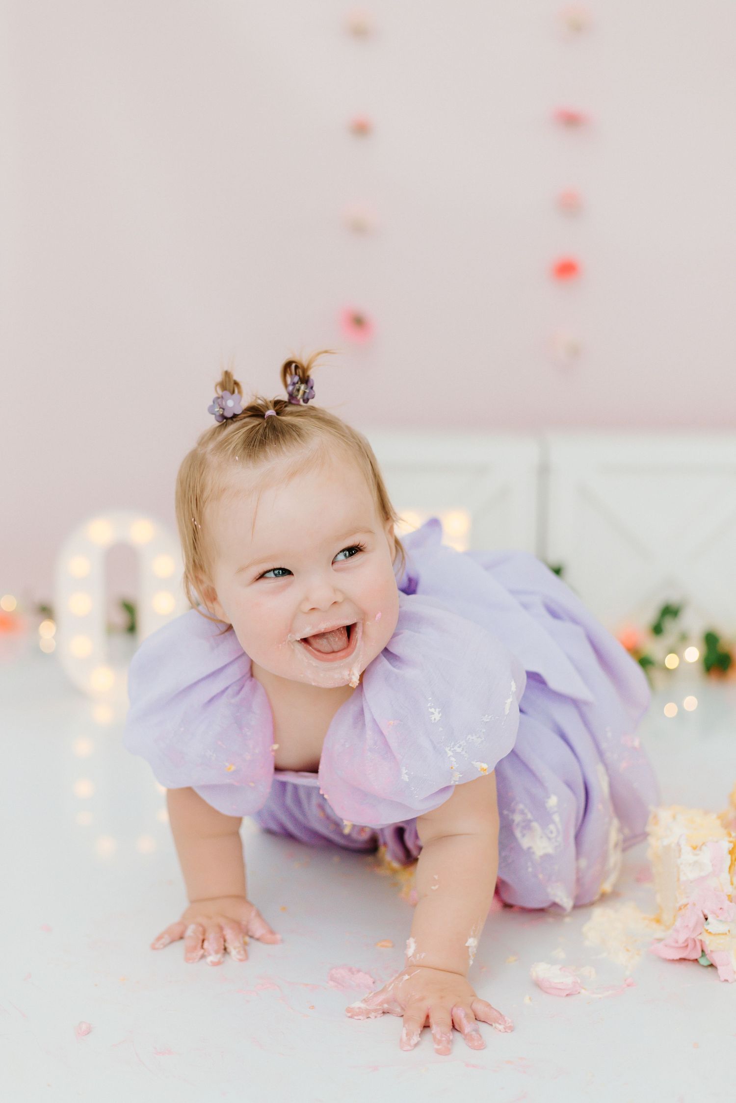 Baby in a purple tulle dress crawls and smiles on white floor with soft lighting and pink decorations.