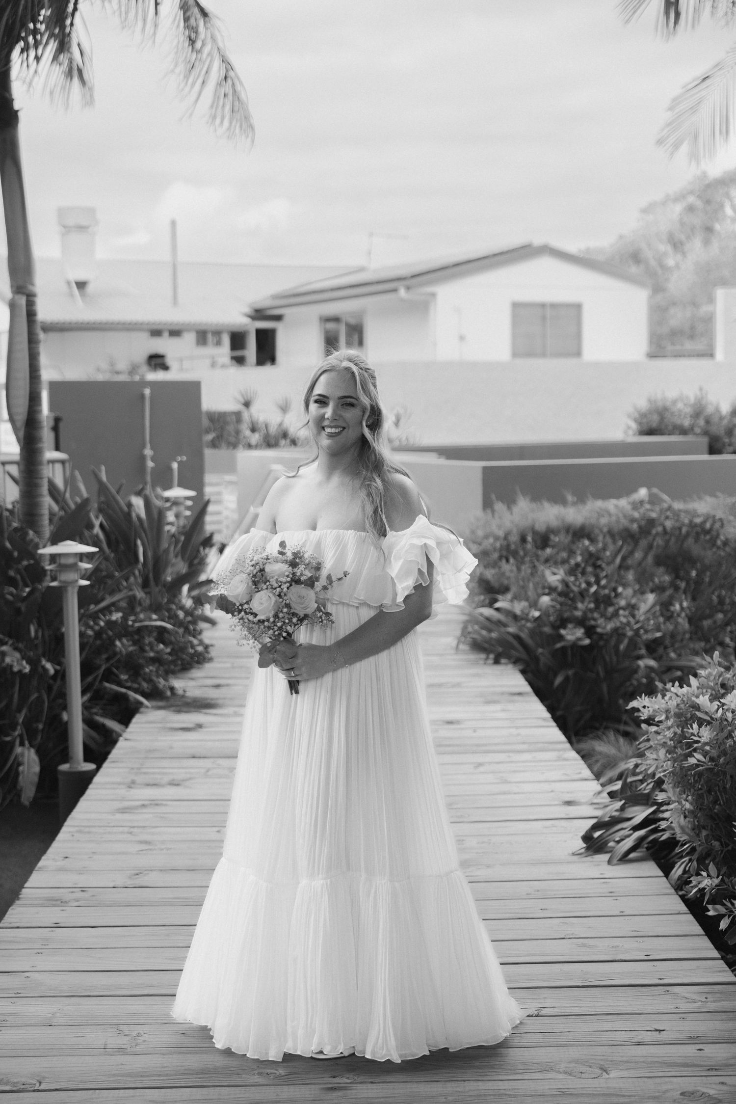Black and white photo series of a bride in a flowing off-shoulder wedding dress walking on a wooden boardwalk.