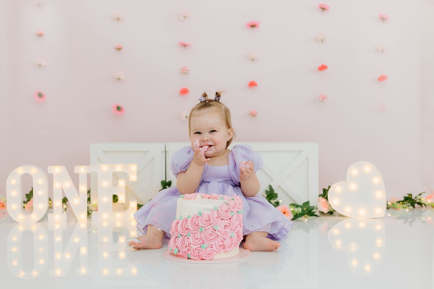 Baby in purple dress celebrates first birthday with pink cake and lit marquee letters spelling ONE in background.