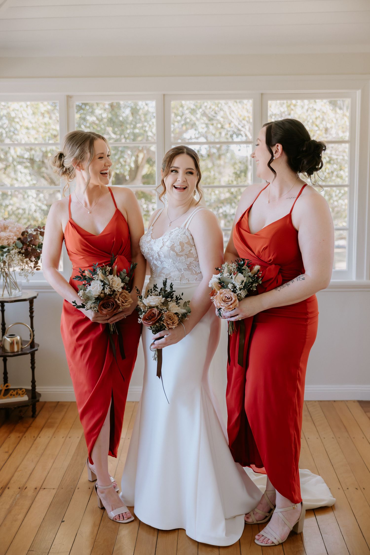 Three women in red and white dresses holding floral bouquets in a bright sunlit room.