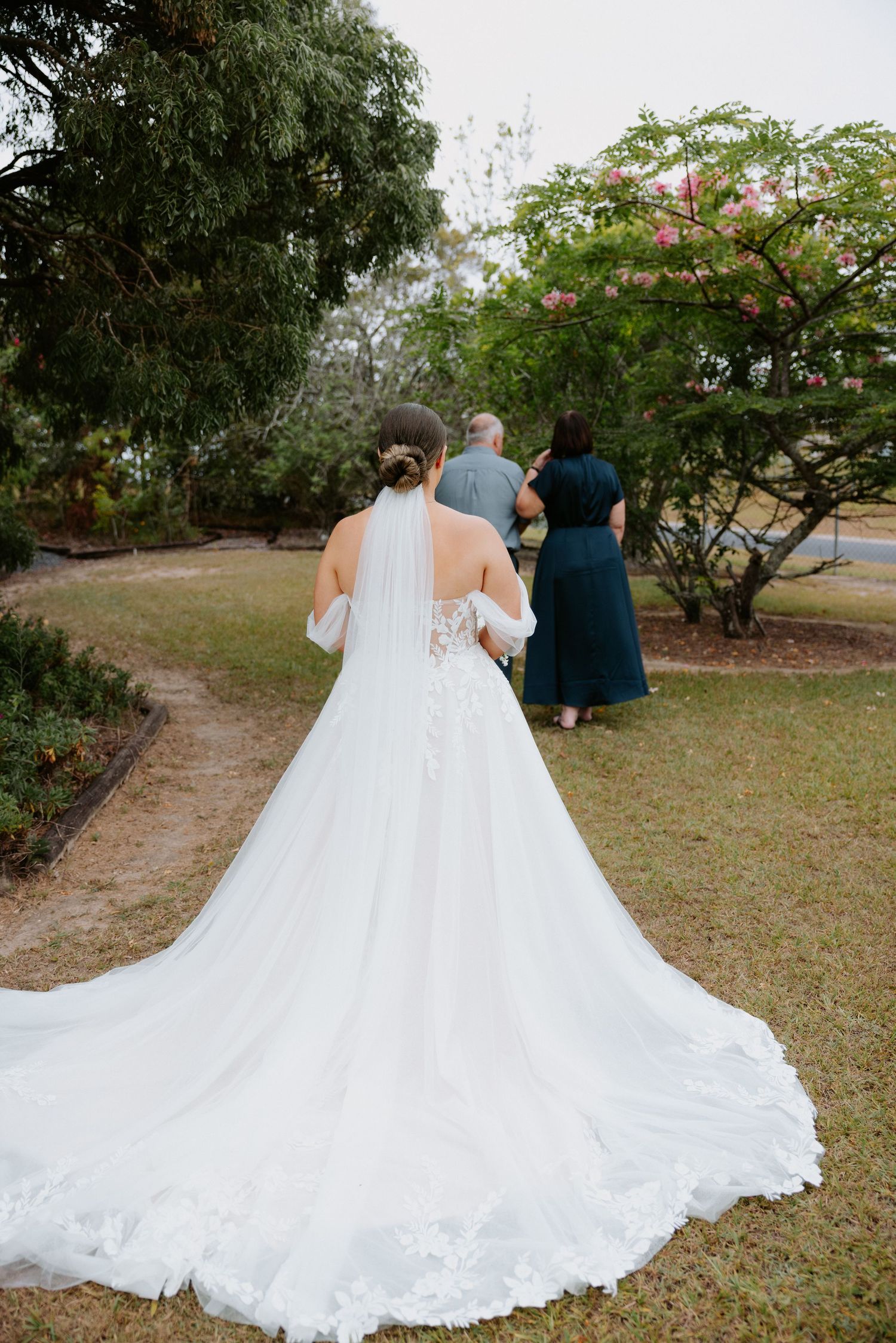 A flowing white wedding dress with a dramatic train trails across a garden path as the couple walks away.