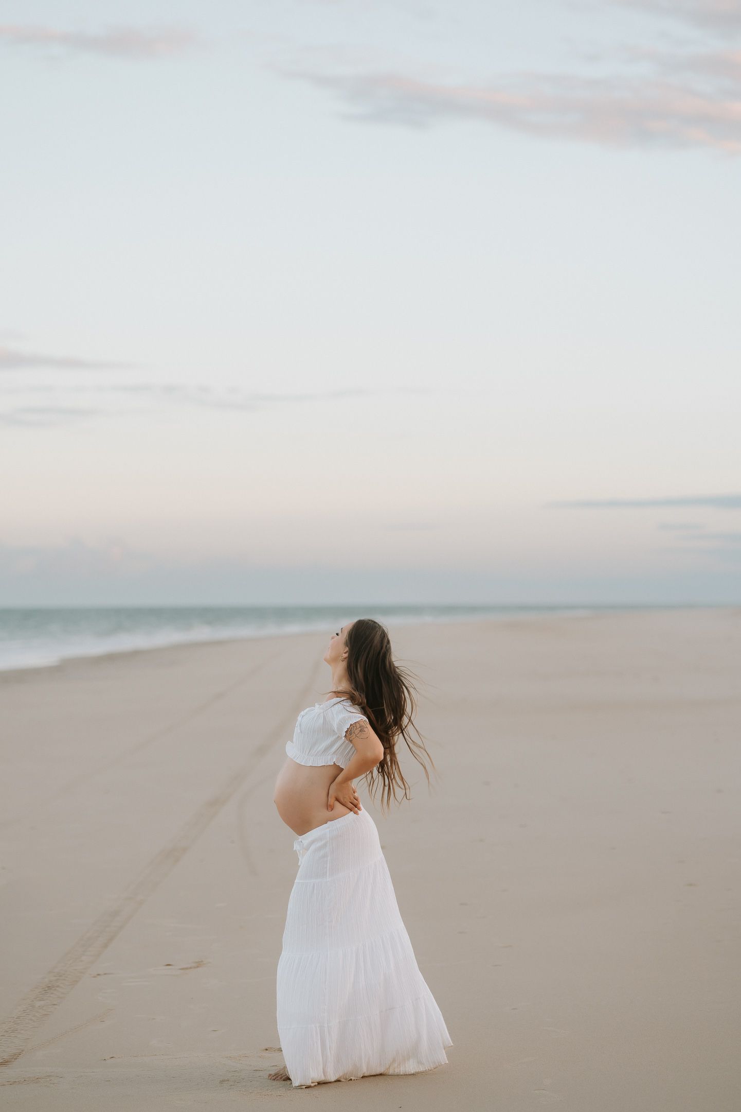 Woman in flowing white dress twirls on empty beach at sunset with cloudy sky in background.