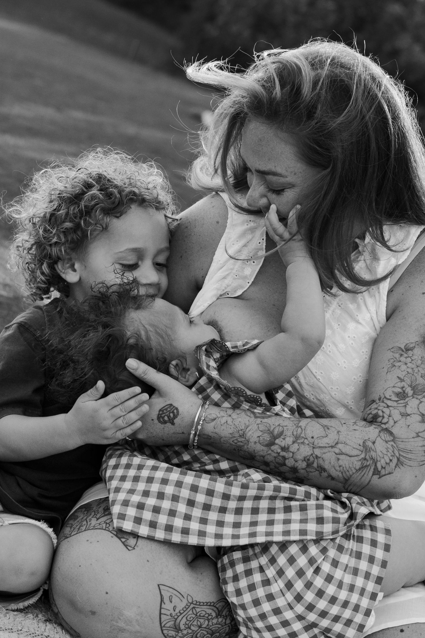 Black and white photo of a tattooed mother breastfeeding while her curly-haired toddler cuddles close outdoors.