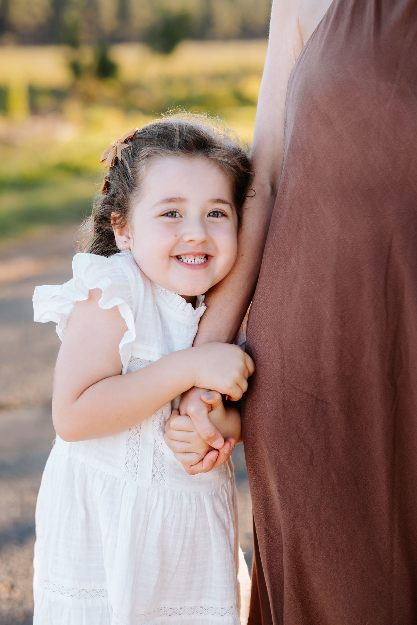 A smiling little girl in a white dress hugs an adult's leg outdoors in a sunny, green setting.