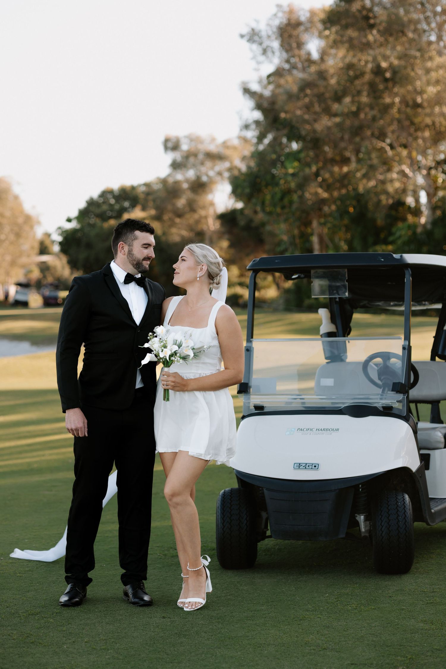 Bride in short white dress holding bouquet stands with groom in black tuxedo beside golf cart on green course.