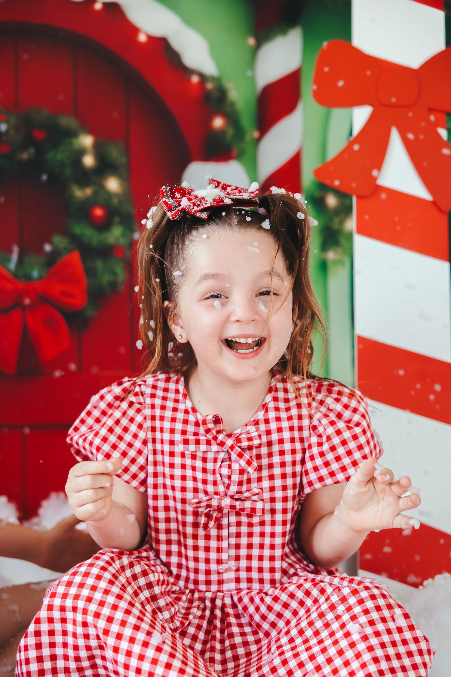 Joyful girl in red gingham dress laughing in Christmas-themed studio with candy canes and falling snow.